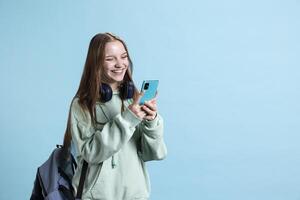 Radiant teenager using smartphone, laughing at amusing social media posts on feed. Joyful person browsing internet websites on phone, amused by funny memes, studio backdrop photo