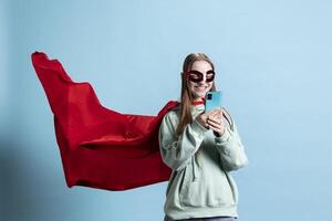 Smiling girl with mask portraying superhero, texting friends on mobile phone, studio background. Happy woman posing as hero in costume with mantle sending messages on smartphone photo