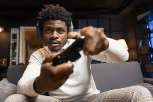 Portrait of gamer playing videogames on console late at night in living room, struggling to stay awake. African american man using gamepad controller to try and finish game level before going to sleep photo