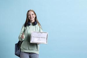 Joyful girl holds school diploma and doing thumbs up hand gesturing, showing approval, having positive mood. Upbeat student with graduation certification doing agreement finger sign, studio background photo