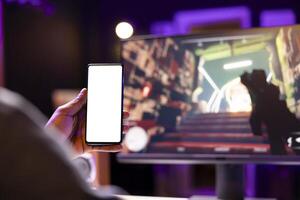 Man texting friends on isolated screen phone while playing first person shooter videogame at home. Gamer looking at mockup smartphone display, enjoying online discussion with mates photo