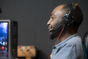 alegre hombre teniendo divertido jugando videojuegos en escritorio computadora, chateando con compañeros de equipo mediante auriculares micrófono relajado jugador comunicado con amigos mediante auriculares micrófono durante juego a hogar foto