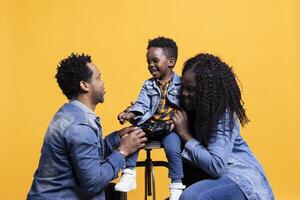Joyful little child playing around with a joystick and sitting next to his parents, posing in front of the camera. African american happy family of three smile against yellow background. photo