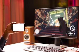 Man watching tutorial on isolated screen phone while playing first person shooter videogame with gun shooting laser bullets. Gamer learning to play game by looking at online guide on mockup cellphone photo