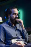 Man enjoying bowl of popcorn while watching cable programs on TV, captivated by scenes. Mesmerized spectator on couch in living room eating snacks and looking at riveting television series photo