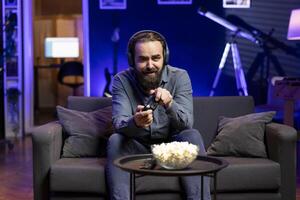 Joyous player at home gaming and enjoying leisure time. Upbeat man sitting on couch in neon lit living room, playing videogame on console, relaxing after hard day at work photo
