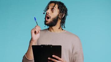 Creative man brainstorming ideas, writing on document using pen. BIPOC photographer using clipboard to complete shoot list for upcoming photography work, isolated over studio background, camera B photo