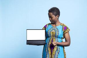 Cheerful woman presenting white laptop display in studio, holding pc that shows isolated mockup layout. Smiling girl in traditional outfit holds pc with blank copyspace screen, advertisement. photo