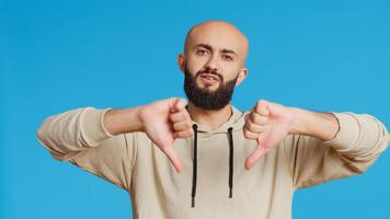 Middle eastern guy showing thumbs down sign on camera, giving dislike symbol and presenting his disappointment. Arab person feeling displeased with negative gesture in studio. Camera 2. photo