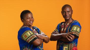 African american people in traditional apparel on camera, feeling carefree and happy with their culture. Joyful married couple standing over orange background, wearing colorful attire. photo