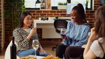 Asian woman chatting with multiracial apartment party guests while eating appetizers. Host enjoying gourmet cheese from charcuterie platter while communicating with friends photo