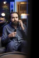 Sad man on couch watching heartbreaking drama movie on streaming service while talking on phone. Person distressed by TV show favorite character dying, discussing with friend on smartphone photo