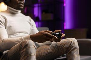 BIPOC man playing intense online videogame in neon illuminated living room, spamming buttons on controller. Gamer participating in internet esports competition, playing on gaming console, close up photo