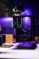 RGB lights illuminated components on open gaming PC tower next to computer peripherals, display and VR headset in empty apartment. Desktop PC in home living room, monitor and virtual reality glasses photo