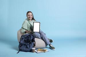 Jolly girl surrounded by school supplies laughing while presenting tablet with isolated screen display, studio background. Joyous student has fun during promotion, showcasing mockup device photo