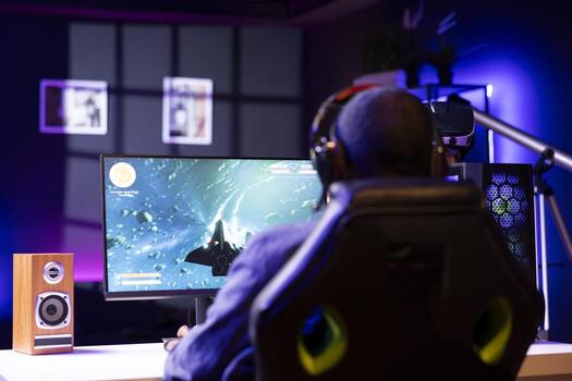 Man wearing headphones playing SF space videogame on PC screen, shooting asteroids. Gamer enjoying spaceship flying game on computer desktop monitor, destroying meteorites photo
