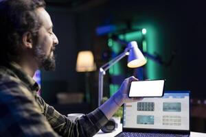 Closeup of a guy grasping a cell phone with a white copyspace screen in front of his laptop. On a smartphone, male individual displays a blank chromakey mockup template while working at home. photo