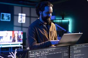Smiling technician doing software debugging using laptop, checking glitches in lines of code displayed on PC monitors. Happy man walking in apartment fixing coding issues using notebook photo