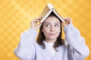 Portrait of happy woman with book on head acting zany, conveying how fun reading is, studio background. Cheerful person wearing novel as hat, promoting literature as entertainment achieving hobby photo