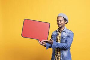 Confident relaxed guy pointing at speech bubble red board in studio, holding a cardboard isolated icon to create a web advertisement. African american man shows empty billboard sign. photo
