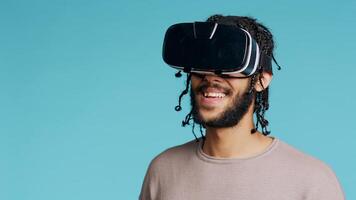 Middle Eastern man wearing virtual reality headset, doing swiping gestures. BIPOC person using high tech futuristic modern VR gadget, isolated over blue studio background, camera B photo