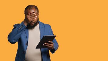 Irked man upset after lacking money while trying to buy stuff online. Grumpy person looking at debit card after getting transaction rejected on tablet, doing facepalm, studio background, camera A photo