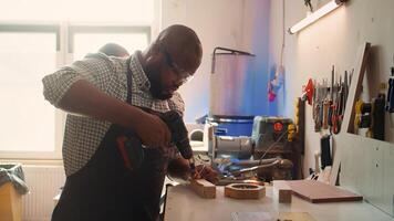African american man in carpentry shop using power drill to create holes for dowels in wooden board. Craftsperson sinks screws into wood block with electric tool, doing precise drilling, camera B photo