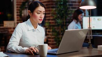 Asian worker sulking in office, having frown on face, feeling annoyed by having too much work to do. Employee writing down info on notepad, gloomily solving tasks late at night photo