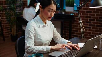 Smiling asian employee writing important emails at computer desk next to full mug of coffee. Cheerful employee typing on laptop, solving tasks, communicating with business partners photo
