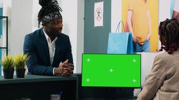 African american employee works with greenscreen on pc at cash register, preparing clothes order to client at checkout. Retail worker using computer with isolated chromakey display. Camera A. photo