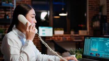 Asian employee taking landline telephone call while working with financial statistics graphs on laptop. Worker talking over the phone and solving tasks in office, doing multitasking, camera A photo
