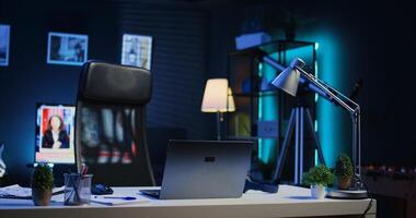 Smart TV left open on news broadcast in empty home office with desk filled with business items. Television set showing VOD channel with Laptop, headphones, plants in blurry foreground, camera B photo