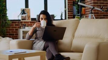 Teleworker lying on couch, drinking coffee while remotely performing tasks in personal office. Freelancer feeling stressed, enjoying hot beverage to relax while inputting data on laptop, camera B photo