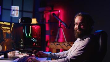 Portrait of smiling man in dark living room playing engaging games on gaming PC at computer desk, chilling after work. Happy gamer contending against foes in online multiplayer shooter photo