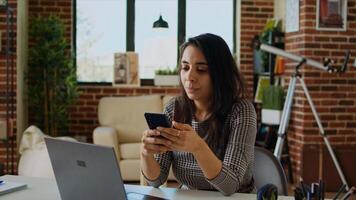 Smiling person checking mobile phone notifications at computer desk, taking break from work. Indian woman in cozy apartment pausing job tasks to read messages on cellphone, camera A zoom in photo
