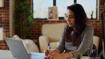 Remote employee at desk doing multitasking, transcribing data from laptop to notepad. Indian woman in apartment trying to complete job tasks before deadline, writing info on paper using pen photo