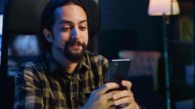 Smiling man at home texting best friend on smartphone, taking break while working in personal office. Cheerful freelancer chatting with mate online using mobile phone, camera A photo