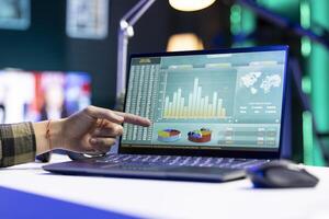 Closeup of a person pointing towards screen displaying financial charts and numbers, representing stock trading. Digital laptop on a desk showing forex trend values and graphs. photo
