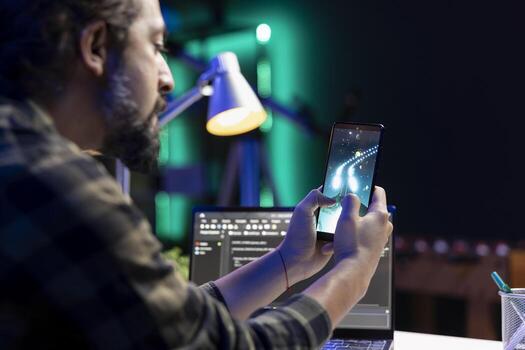 Male programmer taking a break from work, using his mobile device for gaming entertainment. Bearded man grasping his smartphone and playing a space shooter game at home. photo