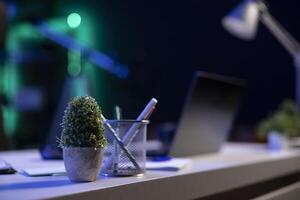 Closeup of various objects placed on a white desk. Different things including a pot of plant, jar of pencils and pens, notebook and laptop are seen in the image. photo