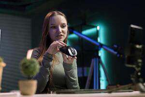 Girl using smartphone on tripod to film for online channel in home studio, presenting wireless mouse to audience. Content creator recording audience computer peripheral device review with phone photo