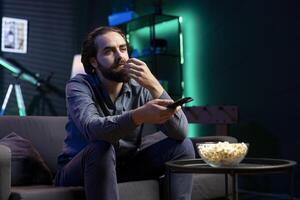 Man in neon lit room on couch eating popcorn while browsing though movies available on streaming service, enjoying time off from work. Viewer consuming snacks at home, holding remote control photo