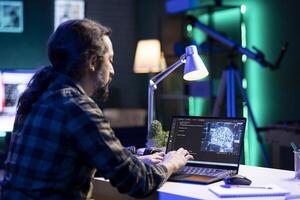 Youthful man using machine learning to manage a complex system on his laptop. Male individual sitting at his desk and typing code to create an online neural network database. photo