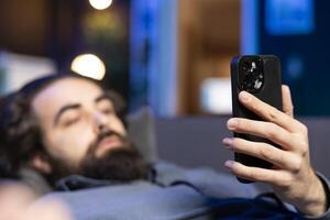 Focus on smartphone with man in blurry background lounged on couch texting friend inviting him to watch movie on streaming service. Close up on phone used by person at home discussing with mate photo