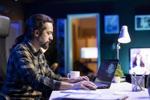 Young man managing a complex system using machine learning on his portable computer. Focused male programmer sitting and typing in code for creating neural network database on laptop. photo