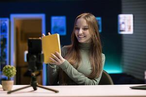 Upbeat gen Z content creator films review of book using smartphone on tripod, presenting synopsis to audience. Media star shows new novel release to subscribers, recording with phone photo