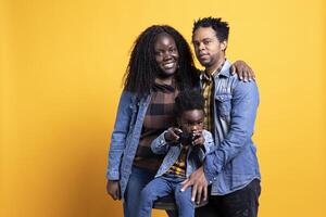 Joyful little child playing with a controller for games, posing with his mother and father in studio. African american family with a toddler standing against yellow background, kid gaming. photo