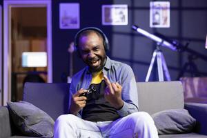 Brass gamer on couch at home insulting rival players while playing competitive multiplayer videogame. Cheeky man throwing middle fingers gesturing at opponents in multiplayer game, playing on console photo