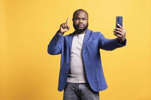 Man using smartphone to take selfies, pointing finger up. BIPOC person taking photos using phone selfie camera, doing hand gesturing, isolated over yellow studio background