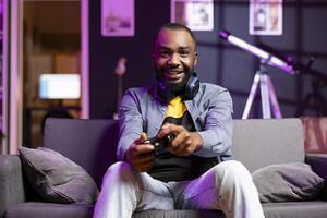 Portrait of cheerful player at home gaming and enjoying leisure time, having fun. Happy man sitting on couch in neon lit apartment, playing videogame on console, relaxing after hard day at work photo